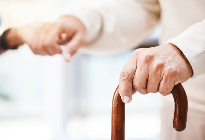 Gehstock mit Hand, die andere Hand wird von einer Pflegerin gehalten